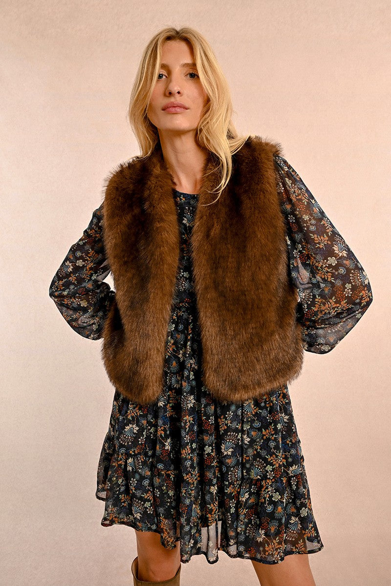 Woman wearing chocolate brown faux fur vest over floral dress against pink background