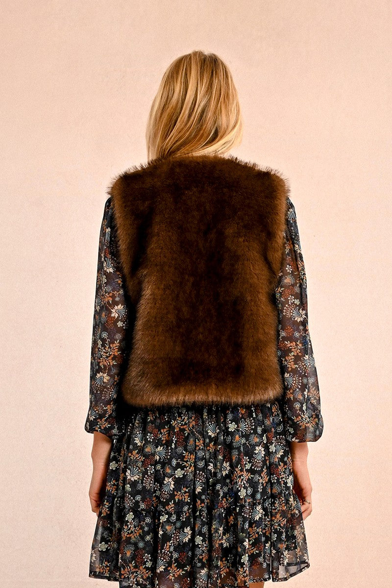 Woman wearing chocolate brown faux fur vest over floral dress against pink background