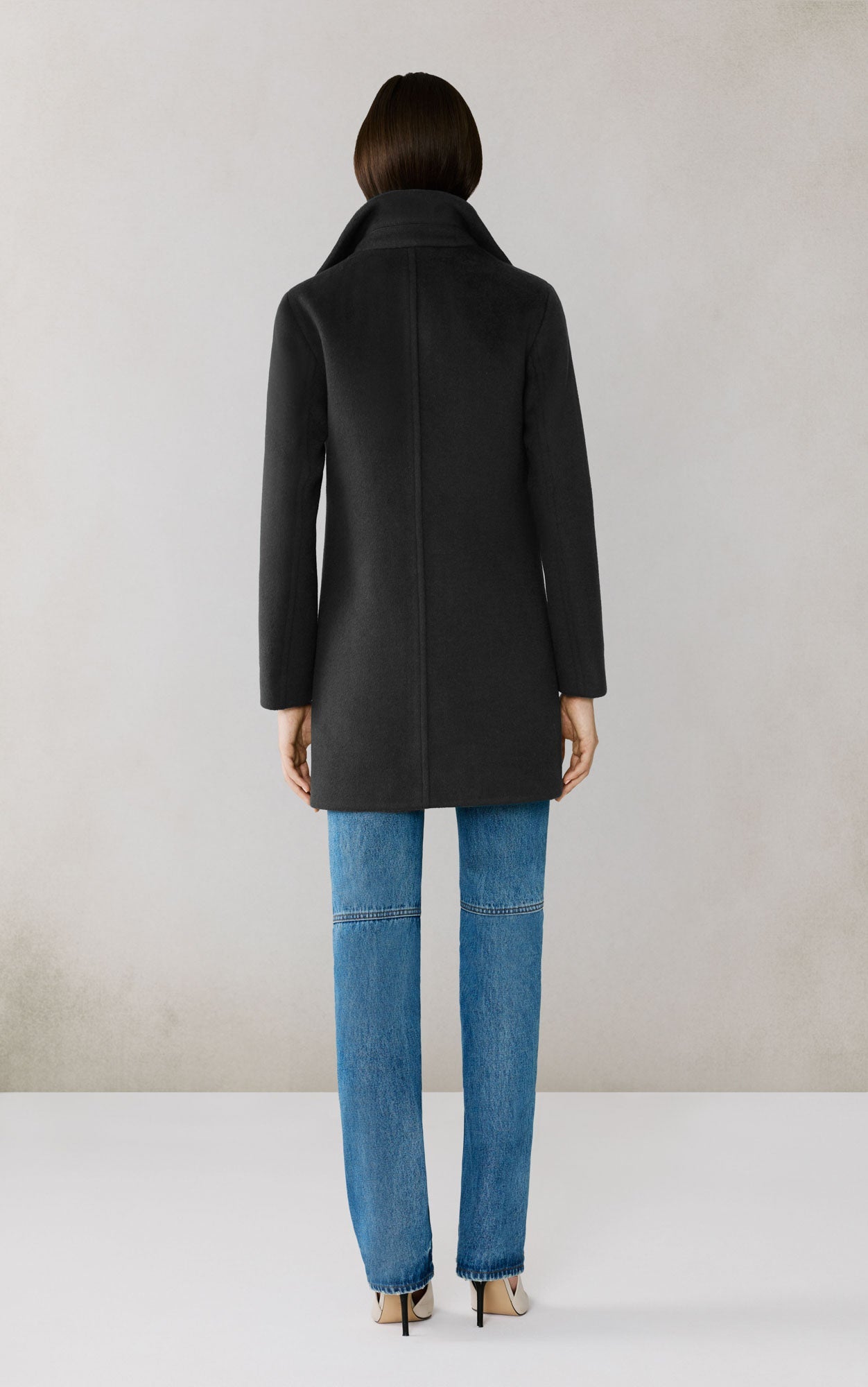Back view of woman wearing black wool coat with blue jeans in studio setting