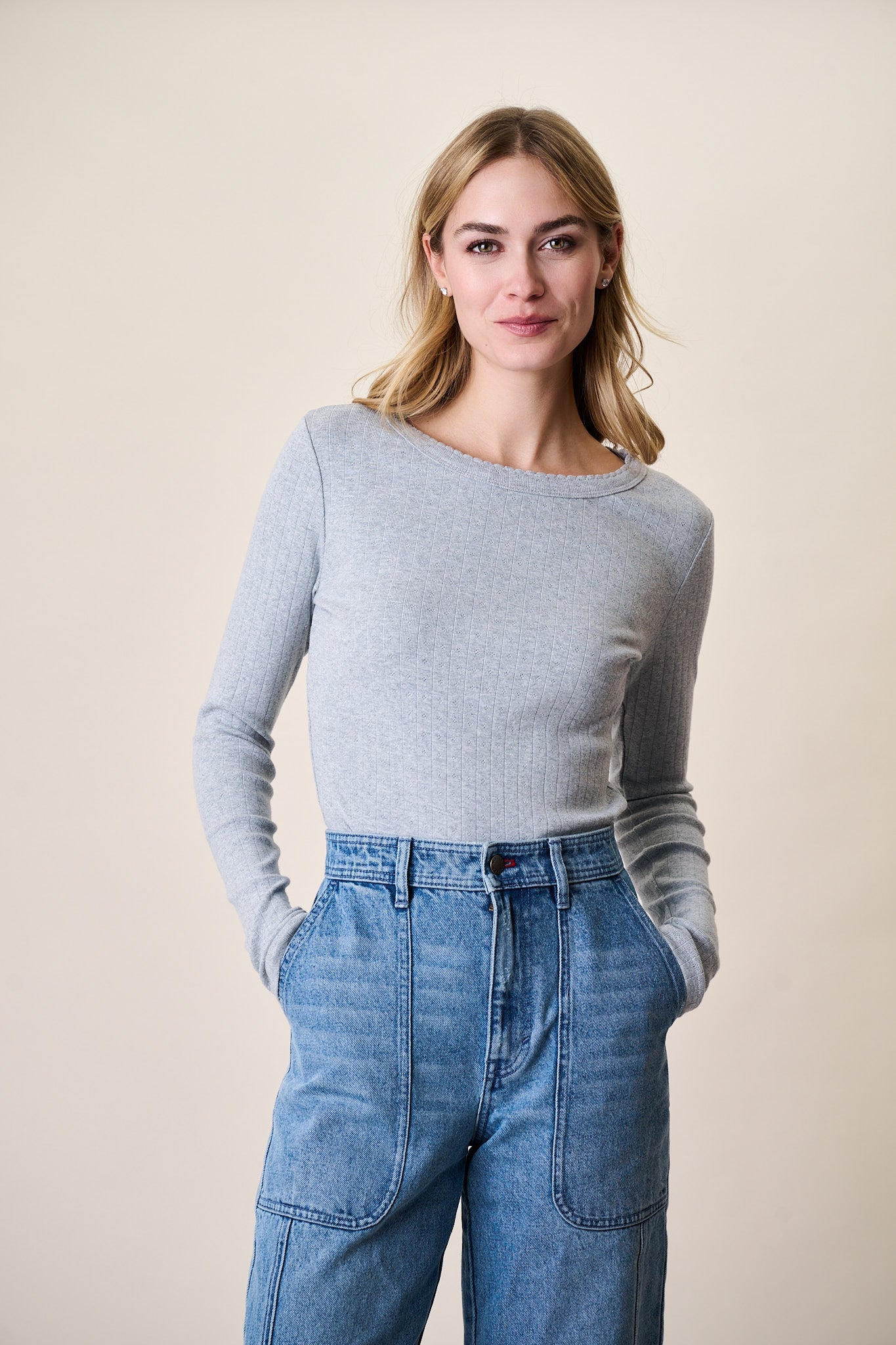 Woman wearing grey pointelle long sleeve crewneck top with blue jeans against beige background