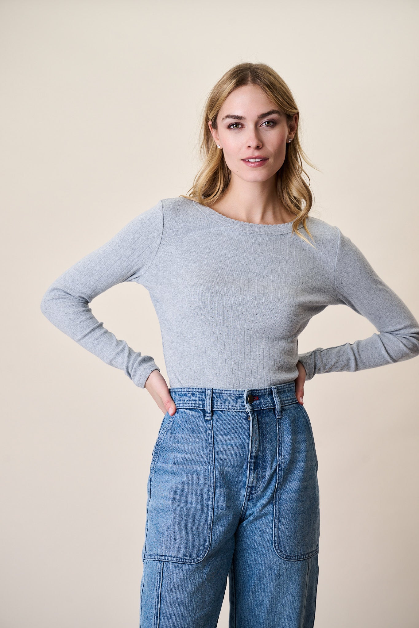Woman wearing grey pointelle long sleeve crewneck sweater with blue jeans against beige background