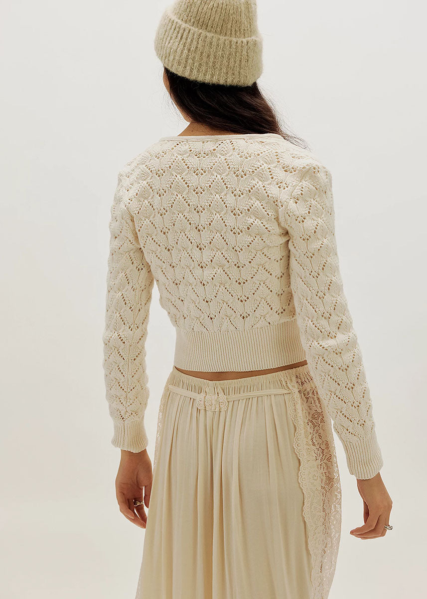 Back view of woman wearing ivory pointelle knit cardigan with matching beanie and pleated skirt