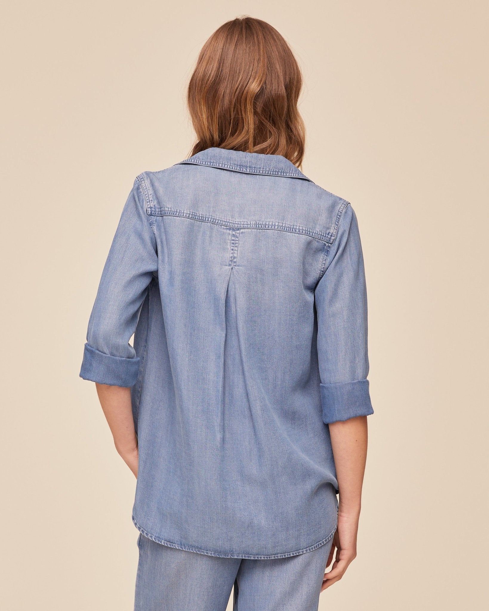 Back view of woman wearing light blue denim button-down shirt with rolled sleeves